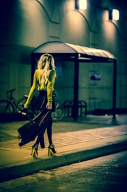 A woman with long blonde hair is walking along a dimly lit street at night, wearing a yellow top, black skirt, and high heels. She carries a large bag and passes a bicycle parked near a bus stop shelter.