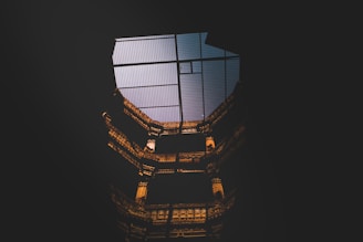 A view of a tall, intricately designed architectural structure, possibly a building interior, with dim lighting accentuating the details. The structure appears to have multiple levels with ornate designs, and the top is partially visible through a mesh-type ceiling.