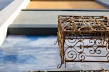 An ornate metal sculpture with intricate scrollwork design casts a shadow on a textured blue and gray surface. The background features geometric shapes in shades of gray and brown, adding a contrasting modern element.