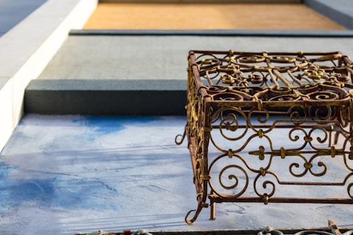 An ornate metal sculpture with intricate scrollwork design casts a shadow on a textured blue and gray surface. The background features geometric shapes in shades of gray and brown, adding a contrasting modern element.