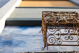 An ornate metal sculpture with intricate scrollwork design casts a shadow on a textured blue and gray surface. The background features geometric shapes in shades of gray and brown, adding a contrasting modern element.