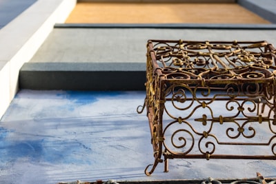 An ornate metal sculpture with intricate scrollwork design casts a shadow on a textured blue and gray surface. The background features geometric shapes in shades of gray and brown, adding a contrasting modern element.