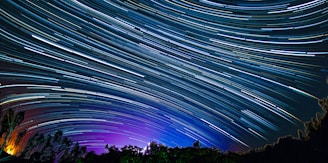A long-exposure photograph capturing the trails of stars moving across a night sky creating dynamic lines of light. The sky transitions from deep shades of blue to purple near the horizon, with silhouettes of trees in the foreground.