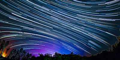 A long-exposure photograph capturing the trails of stars moving across a night sky creating dynamic lines of light. The sky transitions from deep shades of blue to purple near the horizon, with silhouettes of trees in the foreground.