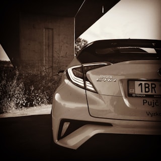 A sepia-toned photograph capturing the rear end of a Toyota C-HR parked under an overpass. The vehicle's contemporary design features angular taillights and a visible license plate with Czech registration. In the background, concrete structures and some foliage are present, contributing to an urban atmosphere.