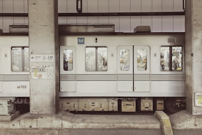 A subway train, with white and gray exterior, is stopped at a station. The train is densely packed with passengers standing inside, visible through the windows. Concrete pillars and signage with Japanese text are present on the platform.