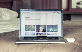 A laptop is open on a bed, displaying a website with nature-themed images. The bedspread is beige, and a wireless mouse sits nearby.