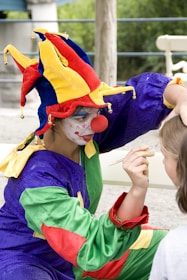 A person dressed as a clown is painting a child's face. The clown is wearing a bright, multicolored jester hat and outfit with blue, red, yellow, and green sections. Their face is painted white with red accents, and they are holding a small paintbrush. The scene appears to take place outdoors in a casual setting.