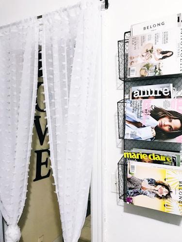 A wall-mounted magazine rack holds several fashion and lifestyle magazines, including 'Belong,' 'Allure,' and 'Marie Claire.' Adjacent to the rack, sheer white curtains with textured polka dots hang partially obscuring a doorway. Large black letters spelling 'LOVE' are partially visible behind the curtain.