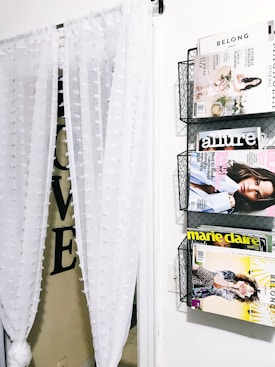 A wall-mounted magazine rack holds several fashion and lifestyle magazines, including 'Belong,' 'Allure,' and 'Marie Claire.' Adjacent to the rack, sheer white curtains with textured polka dots hang partially obscuring a doorway. Large black letters spelling 'LOVE' are partially visible behind the curtain.