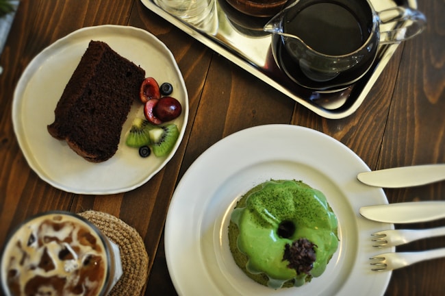 A table set with various desserts and beverages. A slice of chocolate cake with cherries, kiwi, and blueberries on a plate, a green glazed donut with a dark topping on another plate, a glass of iced coffee with cream swirls, and a pot of black coffee on a silver tray.