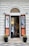 A shop entrance with an arched doorway and mannequins displaying fashionable clothing. The storefront features two large posters on either side with fashion models and the brand name. There are two potted plants placed symmetrically at the entrance.