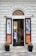 A shop entrance with an arched doorway and mannequins displaying fashionable clothing. The storefront features two large posters on either side with fashion models and the brand name. There are two potted plants placed symmetrically at the entrance.