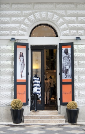 A shop entrance with an arched doorway and mannequins displaying fashionable clothing. The storefront features two large posters on either side with fashion models and the brand name. There are two potted plants placed symmetrically at the entrance.