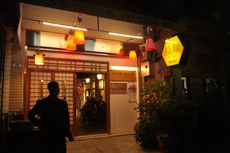 A ramen shop exterior illuminated by warm orange and yellow lighting. The entrance features sliding wooden doors with paper panels. A person stands silhouetted in front of the building, likely looking inside. The sign above the entrance and another hanging sign display the word 'Ramen' in both English and another language. Various light fixtures hang outside, contributing to a cozy and inviting atmosphere.