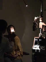 A person is seated in a dark room with one bright light shining on their face. Another individual, likely a medical professional, is examining the seated person's eye using an ophthalmoscope. Medical equipment, including a slit lamp, is visible in the background.