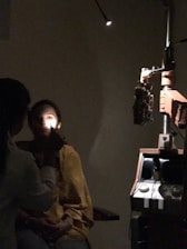 A person is seated in a dark room with one bright light shining on their face. Another individual, likely a medical professional, is examining the seated person's eye using an ophthalmoscope. Medical equipment, including a slit lamp, is visible in the background.