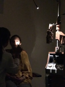 A person is seated in a dark room with one bright light shining on their face. Another individual, likely a medical professional, is examining the seated person's eye using an ophthalmoscope. Medical equipment, including a slit lamp, is visible in the background.