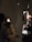 A person is seated in a dark room with one bright light shining on their face. Another individual, likely a medical professional, is examining the seated person's eye using an ophthalmoscope. Medical equipment, including a slit lamp, is visible in the background.