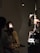 A person is seated in a dark room with one bright light shining on their face. Another individual, likely a medical professional, is examining the seated person's eye using an ophthalmoscope. Medical equipment, including a slit lamp, is visible in the background.