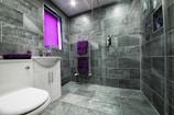 A modern bathroom featuring large gray stone tiles covering the walls and floor. The space includes a white sink with a cabinet and chrome fixtures, accompanied by purple towels on a heated towel rail and a matching soap dispenser. A window with a bright purple blind adds a pop of color. The shower area is equipped with a handheld showerhead and built-in shelving.
