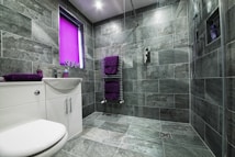 A modern bathroom featuring large gray stone tiles covering the walls and floor. The space includes a white sink with a cabinet and chrome fixtures, accompanied by purple towels on a heated towel rail and a matching soap dispenser. A window with a bright purple blind adds a pop of color. The shower area is equipped with a handheld showerhead and built-in shelving.