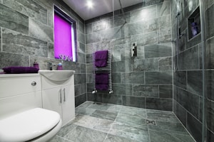 A modern bathroom featuring large gray stone tiles covering the walls and floor. The space includes a white sink with a cabinet and chrome fixtures, accompanied by purple towels on a heated towel rail and a matching soap dispenser. A window with a bright purple blind adds a pop of color. The shower area is equipped with a handheld showerhead and built-in shelving.