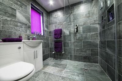 A modern bathroom featuring large gray stone tiles covering the walls and floor. The space includes a white sink with a cabinet and chrome fixtures, accompanied by purple towels on a heated towel rail and a matching soap dispenser. A window with a bright purple blind adds a pop of color. The shower area is equipped with a handheld showerhead and built-in shelving.