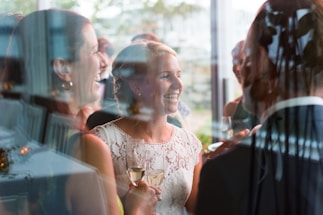 A group of people is gathered at a social event, with a focus on a woman in a white lace outfit holding a glass of champagne. They appear to be laughing and conversing in a joyful manner. The scene is reflected through a window, adding depth and a sense of intimacy to the moment.