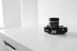 A vintage camera sits on a clean white surface. The composition is minimalist with a focus on the camera, which has intricate dials and a lens. The background is softly lit and blurred, emphasizing the sleek design of the camera.