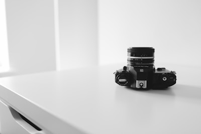 A vintage camera sits on a clean white surface. The composition is minimalist with a focus on the camera, which has intricate dials and a lens. The background is softly lit and blurred, emphasizing the sleek design of the camera.