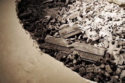 A large pile of objects including electronics and mechanical parts is embedded in a concrete surface. The items appear to be deliberately arranged, creating a dense and chaotic scene. The lighting casts a dramatic shadow over the collection, highlighting keyboards, radios, wheels, and other miscellaneous components.