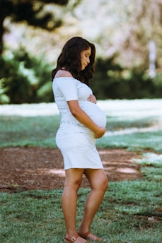 A pregnant woman is standing outdoors on grass, wearing a striped dress and sandals. She is gently holding her belly with both hands, and her expression is serene. The background consists of blurred greenery, suggesting a park or garden setting.