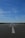 A wide, open road stretches into the distance under a clear blue sky. A large white arrow is painted on the asphalt, pointing forward. Several people are walking or cycling along the road. The horizon is visible with some industrial buildings and greenery in the distance.
