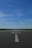 A wide, open road stretches into the distance under a clear blue sky. A large white arrow is painted on the asphalt, pointing forward. Several people are walking or cycling along the road. The horizon is visible with some industrial buildings and greenery in the distance.