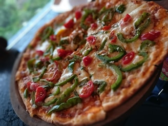 A freshly baked pizza topped with vibrant green bell peppers and juicy red cherry tomatoes. The crust appears golden and crispy, and the cheese is melted perfectly, creating a mouth-watering appearance. The pizza is served on a wooden board, and the blurred background suggests an outdoor or casual dining setting.