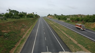 A wide highway stretches into the distance, dividing into two main lanes in each direction. The road is bordered on both sides by grassy embankments and scattered trees, suggesting a rural setting. A colorful truck is moving along the right side of the road, with a few smaller vehicles further ahead.
