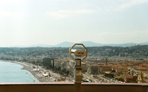 A scenic view of a seaside city with a prominent coastline and a mountain range in the background. In the foreground, there is a mounted binocular viewer overlooking the city and beach, suggesting a popular tourist viewpoint.