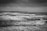 Black and white photograph of a coastal landscape with crashing waves against rows of wooden posts protruding from the water. The sky is overcast, contributing to a somber and dramatic atmosphere. The horizon shows a distant landmass, adding depth to the scene.