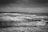 Black and white photograph of a coastal landscape with crashing waves against rows of wooden posts protruding from the water. The sky is overcast, contributing to a somber and dramatic atmosphere. The horizon shows a distant landmass, adding depth to the scene.