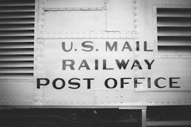 The image features a vintage railway car with the words 'U.S. Mail Railway Post Office' prominently displayed. The surface is metallic and riveted, suggesting an industrial design from the early to mid-20th century. The image is in black and white, enhancing its historical feel.