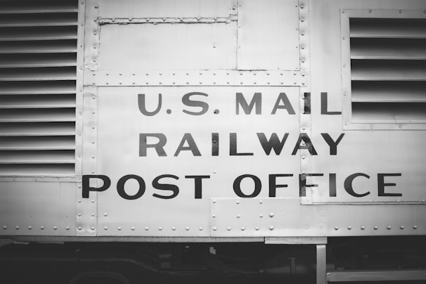 The image features a vintage railway car with the words 'U.S. Mail Railway Post Office' prominently displayed. The surface is metallic and riveted, suggesting an industrial design from the early to mid-20th century. The image is in black and white, enhancing its historical feel.