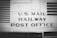 The image features a vintage railway car with the words 'U.S. Mail Railway Post Office' prominently displayed. The surface is metallic and riveted, suggesting an industrial design from the early to mid-20th century. The image is in black and white, enhancing its historical feel.