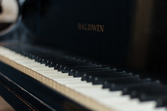 A close-up view of a piano keyboard with the brand name Baldwin visible on the piano body. The keys have a mix of black and white keys with visible shadows.
