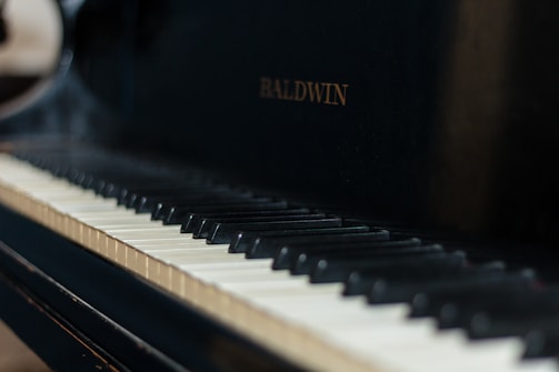 A close-up view of a piano keyboard with the brand name Baldwin visible on the piano body. The keys have a mix of black and white keys with visible shadows.