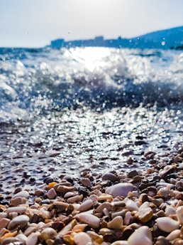 Waves approach a pebbly shore in the foreground with sunlight reflecting off the water. The background features blurred silhouette of distant buildings and hills under a blue sky.