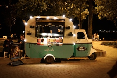 A small food truck serving waffles is illuminated with white lights. It is parked on a cobblestone path near a park with trees. The truck is branded with 'Waffle Love' and has a turquoise and white color scheme. There are two people sitting nearby, eating, and a poster advertising waffle sandwiches.