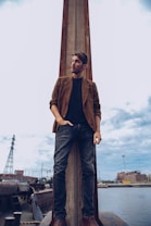 A man stands confidently with one hand in his pocket and the other resting by his side. He is positioned in front of a tall, industrial-looking structure near a body of water, with buildings and industrial elements visible in the background. The sky is overcast, and the scene evokes an urban, gritty atmosphere.