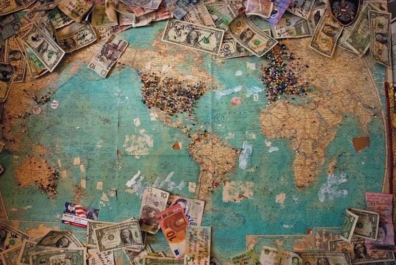 A vintage world map covered with various international currency notes serves as a background. Colorful pins are densely clustered over some regions, indicating highlighted areas. The currencies range from dollars to euros and others, scattered across the edges, giving a sense of travel or exploration.