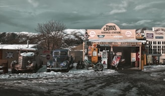A vintage service station scene, featuring an old gas station with classic cars parked in front. Snow is scattered on the ground, and a bicycle leans against the station. Various signs and logos, like Shell and BP, decorate the building, and a stand-alone gas pump is visible. The background shows a tree without leaves, and mountains under a cloudy sky.